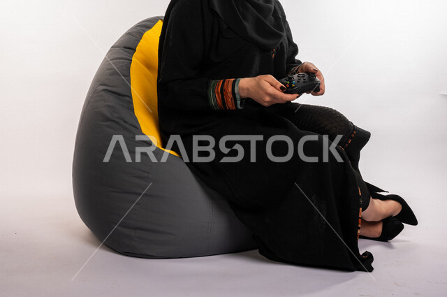 بورتريه لإمرأه عربيه سعوديه خليجيه تجلس على أريكه مريحه, اللعب بألعاب البلايستيشن,  استخدام يد التحكم عن بعد, ايماءات وجه ويدين تدل على التفاعل والإندماج مع اللعبه, تسليه وترفيه, قضاء وقت ممتع مع الألعاب الاكترونيه