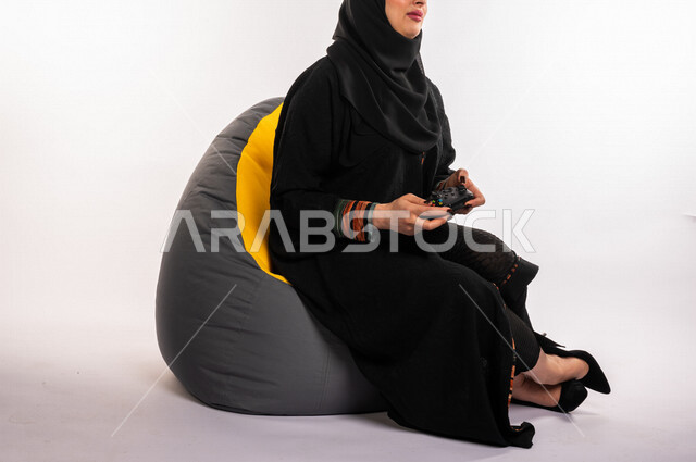 بورتريه لإمرأه عربيه سعوديه خليجيه تجلس على أريكه مريحه, اللعب بألعاب البلايستيشن,  استخدام يد التحكم عن بعد, ايماءات وجه ويدين تدل على التفاعل والإندماج مع اللعبه, تسليه وترفيه, قضاء وقت ممتع مع الألعاب الاكترونيه