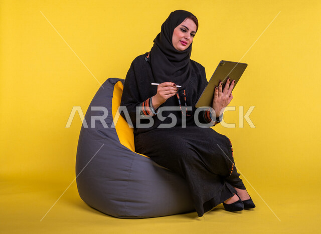 A Saudi Arabian Gulf woman sitting on a comfortable sofa, completing work tasks via the tablet, managing projects remotely, playing on the mobile device, making video calls over the Internet, watching a movie, having fun, having a good time, yellow backgr