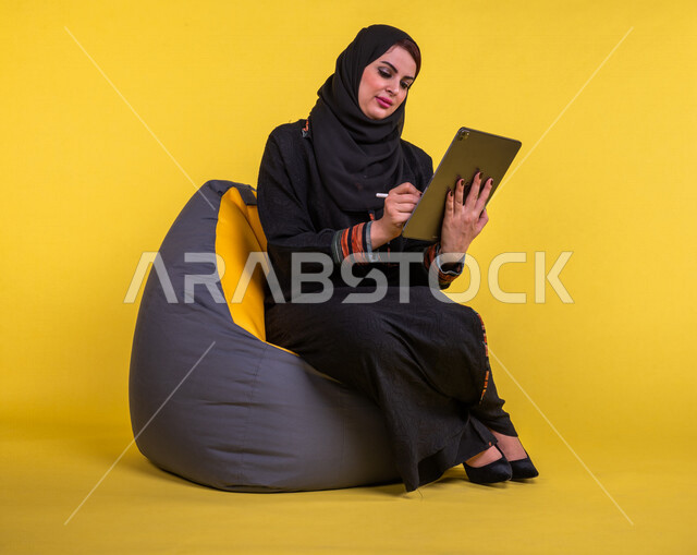 إمرأه عربيه سعوديه خليجيه تجلس على أريكه مريحه, انجاز مهام العمل عبر التابلت, إدارة المشاريع عن بعد, اللعب على الجهاز المحمول, إجراء مكالمات فيديو عبر الإنترنت, مشاهدة فلم, تسليه وترفيه, قضاء وقت ممتع, خلفيه صفراء