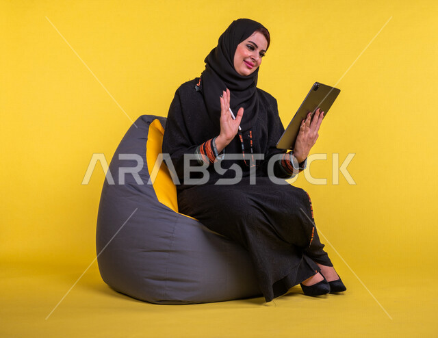 إمرأه عربيه سعوديه خليجيه تجلس على أريكه مريحه, انجاز مهام العمل عبر التابلت, إدارة المشاريع عن بعد, اللعب على الجهاز المحمول, إجراء مكالمات فيديو عبر الإنترنت, مشاهدة فلم, تسليه وترفيه, قضاء وقت ممتع, خلفيه صفراء