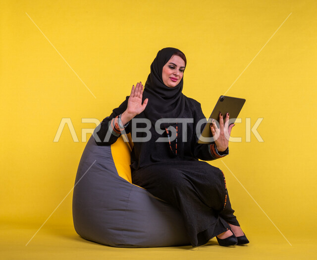 إمرأه عربيه سعوديه خليجيه تجلس على أريكه مريحه, انجاز مهام العمل عبر التابلت, إدارة المشاريع عن بعد, اللعب على الجهاز المحمول, إجراء مكالمات فيديو عبر الإنترنت, مشاهدة فلم, تسليه وترفيه, قضاء وقت ممتع, خلفيه صفراء