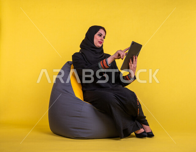 A Saudi Arabian Gulf woman sitting on a comfortable sofa, completing work tasks via the tablet, managing projects remotely, playing on the mobile device, making video calls over the Internet, watching a movie, having fun, having a good time, yellow backgr