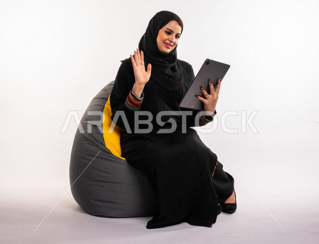 Portrait of a Saudi Arabian Gulf woman sitting on a comfortable sofa, accomplishing work tasks via the tablet, managing projects remotely, playing on the mobile device, making video calls over the Internet, watching a movie, having fun and having a good t