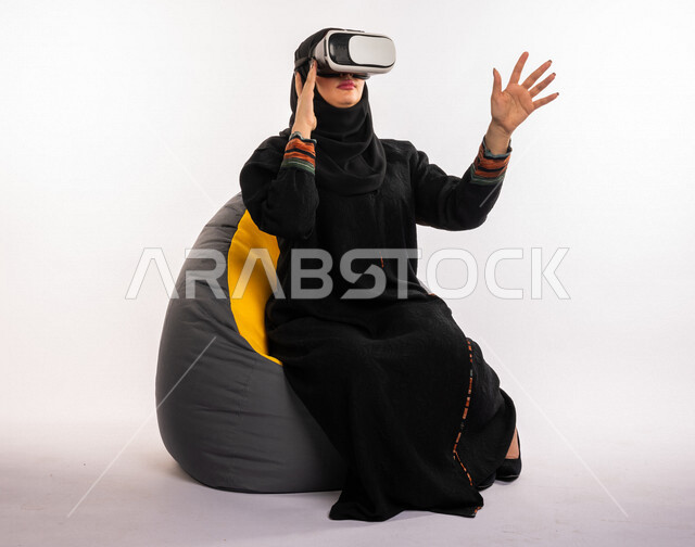 بورتريه لإمرأه عربيه سعوديه خليجيه تجلس على أريكه مريحه, إرتداء نظارة الواقع الإفتراضي, إيماءات وجه ويدين تدل على الإندماج مع العالم الإفتراضي ثلاثي الأبعاد, تسليه وترفيه, قضاء وقت ممتع, تطور التقنيه والتكنولوجيا