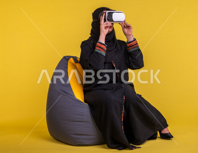 إمرأه عربيه سعوديه خليجيه تجلس على أريكه مريحه, إرتداء نظارة الواقع الإفتراضي, إيماءات وجه ويدين تدل على الإندماج مع العالم الإفتراضي ثلاثي الأبعاد, تسليه وترفيه, قضاء وقت ممتع, تطور التقنيه والتكنولوجيا, خلفيه صفراء