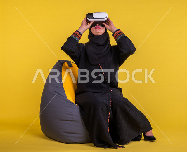 إمرأه عربيه سعوديه خليجيه تجلس على أريكه مريحه, إرتداء نظارة الواقع الإفتراضي, إيماءات وجه ويدين تدل على الإندماج مع العالم الإفتراضي ثلاثي الأبعاد, تسليه وترفيه, قضاء وقت ممتع, تطور التقنيه والتكنولوجيا, خلفيه صفراء