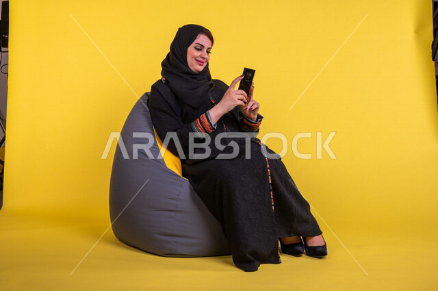 بورتريه لسيدة عربية خليجية سعودية مبتسمة تستخدم الهاتف المحمول ، استخدام مواقع التواصل الاجتماعي ، اجراء مكالمات الفيديو ، التسويق الالكتروني ، تصفح الانترنت ، العمل عن بعد ، خلفية صفراء