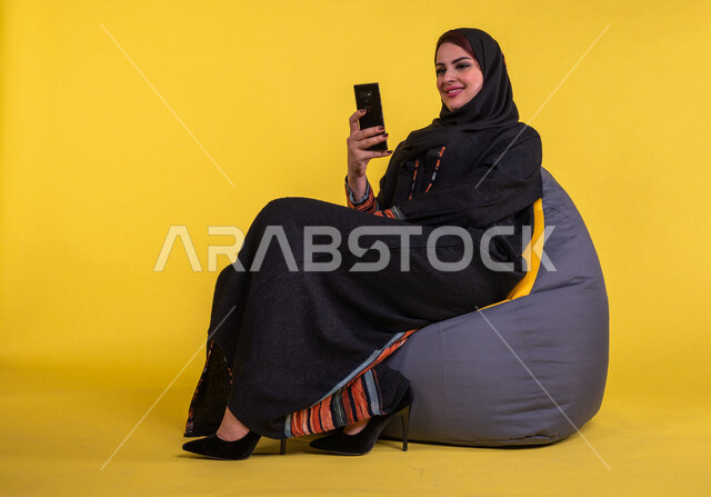 بورتريه لسيدة عربية خليجية سعودية مبتسمة تستخدم الهاتف المحمول ، استخدام مواقع التواصل الاجتماعي ، اجراء مكالمات الفيديو ، التسويق الالكتروني ، تصفح الانترنت ، العمل عن بعد ، خلفية صفراء