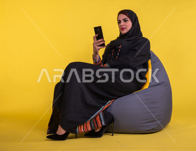 Portrait of a Saudi Arabian Gulf woman using a mobile phone, using social networking sites, making video calls, e-marketing, surfing the Internet, working remotely, yellow background