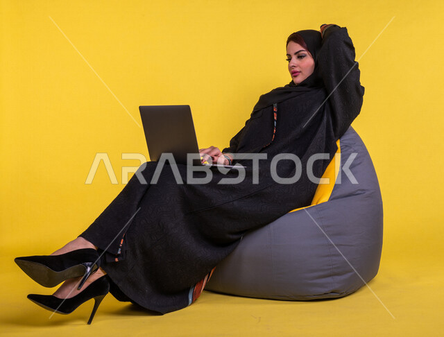 بورتريه لسيدة عربية خليجية سعودية تستخدم الحاسوب المحمول لانجاز الاعمال عن بعد ، اجراء مكالمات الفيديو ، مشاهدة البرامج والافلام ،  التسويق الالكتروني ، تصفح الانترنت ،  خلفية صفراء