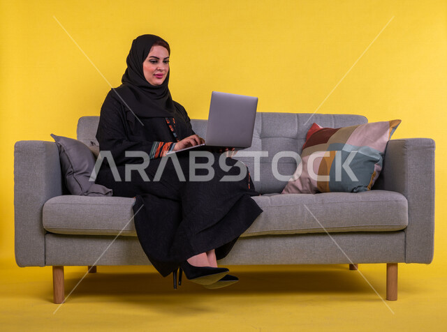 بورتريه لسيدة عربية خليجية سعودية تستخدم الحاسوب المحمول لانجاز الاعمال عن بعد ، اجراء مكالمات الفيديو ، مشاهدة البرامج والافلام ،  التسويق الالكتروني ، تصفح الانترنت ،  خلفية صفراء