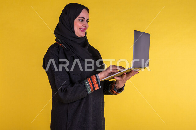 بورتريه لسيدة عربية خليجية سعودية مبتسمة تستخدم الحاسوب المحمول لانجاز الاعمال عن بعد ، اجراء مكالمات الفيديو ، مشاهدة البرامج والافلام ، التسويق الالكتروني ، تصفح الانترنت ، خلفية صفراء