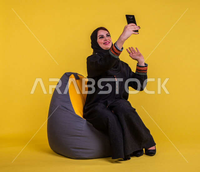 Portrait of a smiling Saudi Arabian Gulf woman using a mobile phone to make video calls, raising her hand to say peace, remote work meeting, communicating with friends, taking souvenir photos, entertainment, yellow background