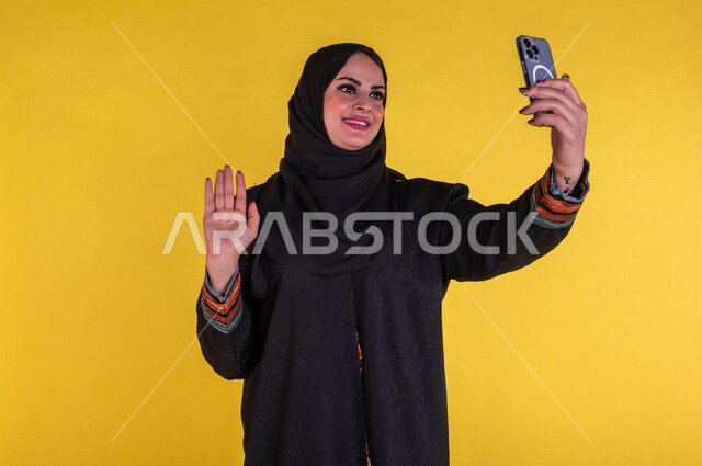 Portrait of a smiling Saudi Arabian Gulf woman using a mobile phone to make video calls, raising her hand to say peace, remote work meeting, communicating with friends, taking souvenir photos, entertainment, yellow background