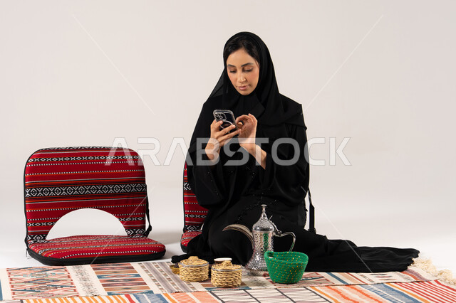 امرأة سعودية بالعباءة تستخدم الجوال مع دلة القهوة