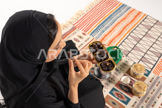 امرأة سعودية بالعباءة تتناول التمر على سجادة تراثية