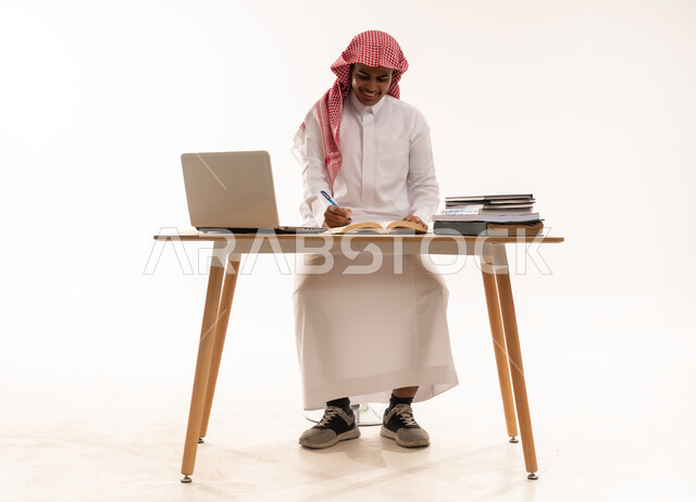 طالب سعودي يكتب على مكتب مع حاسوب محمول وكتب دراسية
