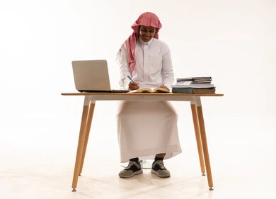 Saudi Student Writing at Desk with Laptop and Books