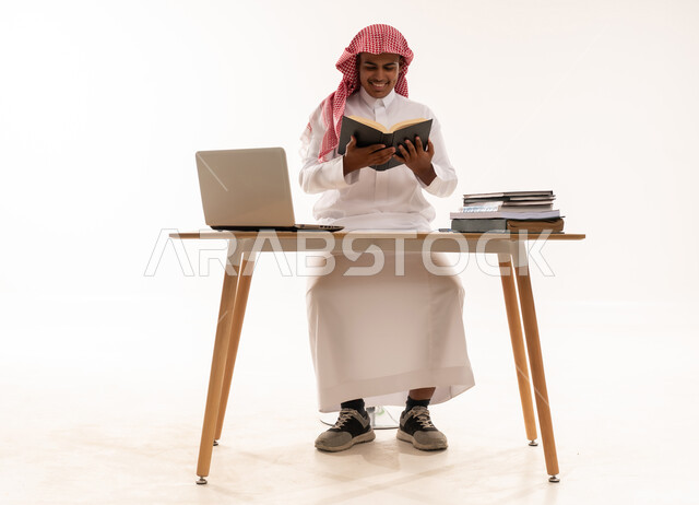 رجل سعودي يقرأ كتاباً على مكتب مع لابتوب