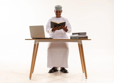 Omani Man Reading Book at Desk with Laptop