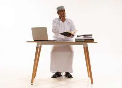 رجل عماني يجلس خلف مكتب مع لابتوب وكتب
