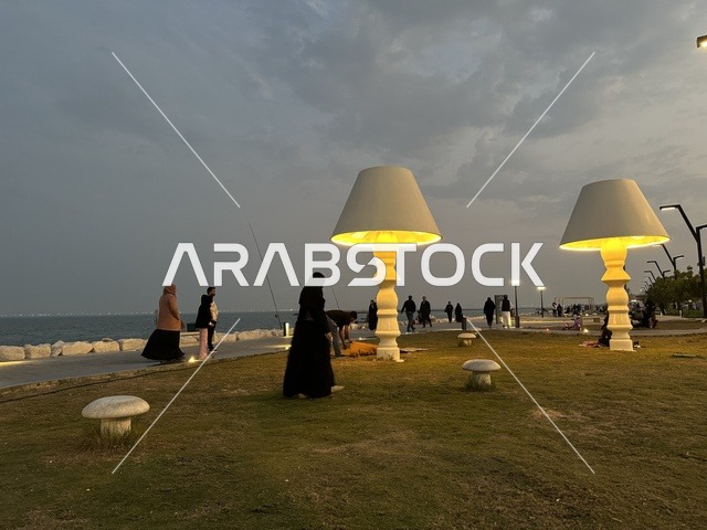 The lights and street lamps at night in the Hams Walkway in the Eastern Province, Saudi Arabia, create a calm family atmosphere, a modern strolling experience, an open entertainment destination, spaces for walking and relaxation, and outdoor seating and benches.