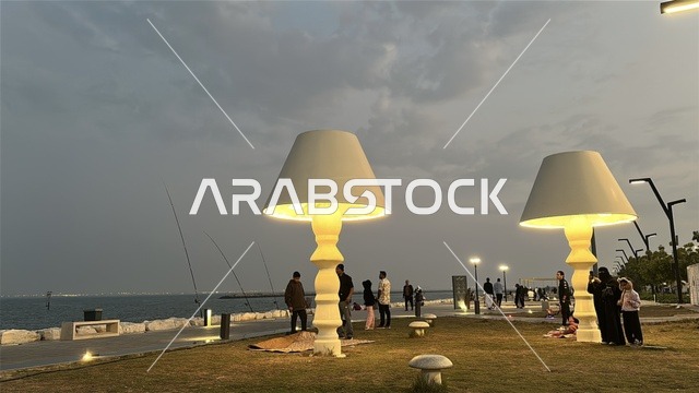 The lights and street lamps at night in the Hams Walkway in the Eastern Province, Saudi Arabia, create a calm family atmosphere, a modern strolling experience, an open entertainment destination, spaces for walking and relaxation, and outdoor seating and benches.