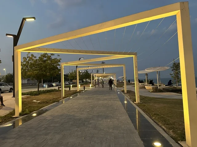 The lights and street lamps at night in the Hams Walkway in the Eastern Province, Saudi Arabia, create a calm family atmosphere, a modern strolling experience, an open entertainment destination, spaces for walking and relaxation, and outdoor seating and benches.