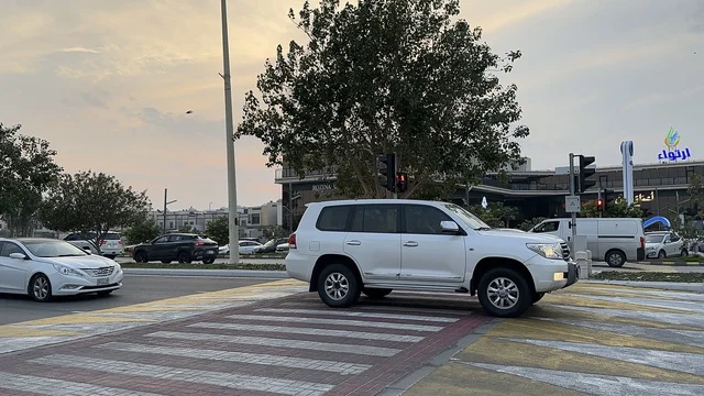 Pedestrian and vehicle crossing at the Hams walkway in the Eastern Province at night, Saudi Arabia, a calm family atmosphere, a modern strolling experience, an open entertainment destination, spaces for walking and relaxing, outdoor seating and benches.