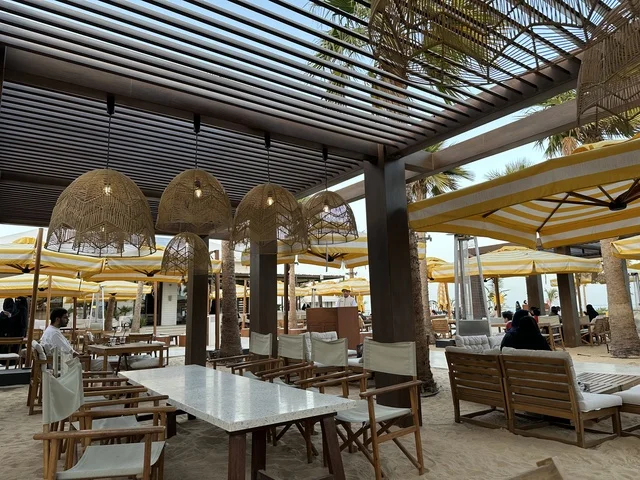 طاولات وكراسي خشبية مرتبة على رمل ناعم، سولت البحر وجهة ترفيهية وشاطئية عائلية تقع على طريق الكورنيش في الخبر في المملكة العربية السعودية، شاطئ رملي مع وجود مظلات وأشجار نخيل، تجديدات إضافية وتطويرات في 3  يناير 2026