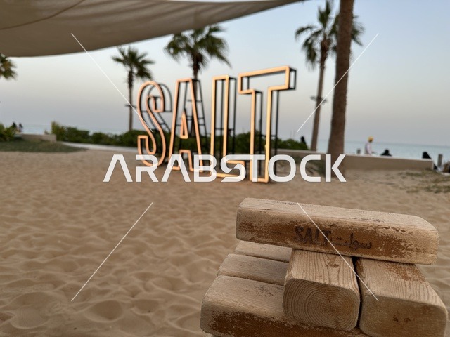 The wooden block in the front bears the name Salt, a sandy beach with umbrellas and palm trees. Salt Beach is a family-friendly recreational and beach destination located on the Corniche Road in Al Khobar, Saudi Arabia, with additional renovations and developments on January 3, 2026.