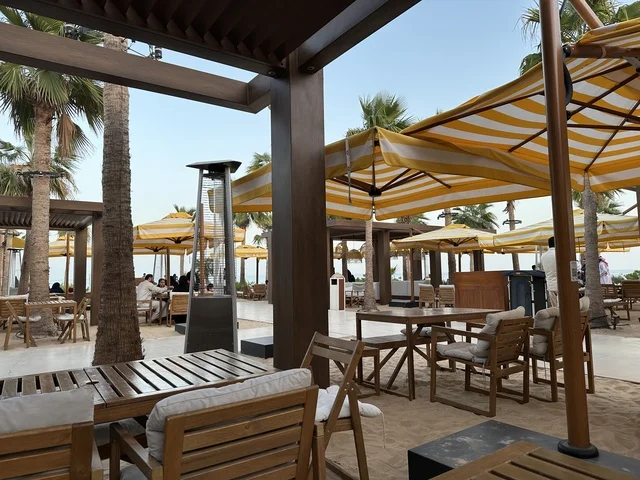 طاولات وكراسي خشبية مرتبة على رمل ناعم، سولت البحر وجهة ترفيهية وشاطئية عائلية تقع على طريق الكورنيش في الخبر في المملكة العربية السعودية، شاطئ رملي مع وجود مظلات وأشجار نخيل، تجديدات إضافية وتطويرات في 3  يناير 2026