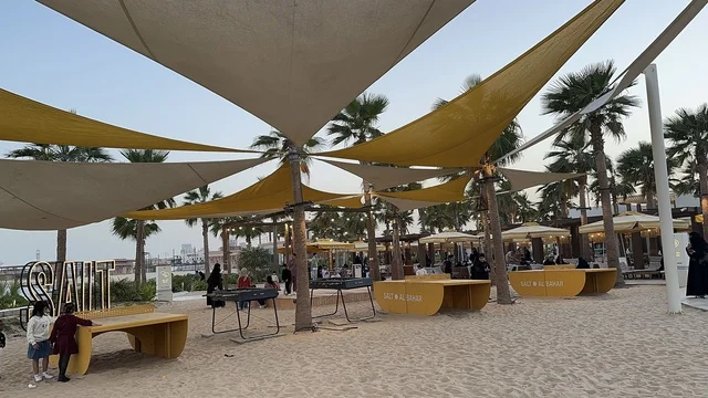 أشجار نخيل أضواء صغيرة حول جذوعها، سولت البحر وجهة ترفيهية وشاطئية عائلية تقع على طريق الكورنيش في الخبر في المملكة العربية السعودية، تجديدات إضافية وتطويرات في 3  يناير 2026، شاطئ رملي مع وجود مظلات وأشجار نخيل