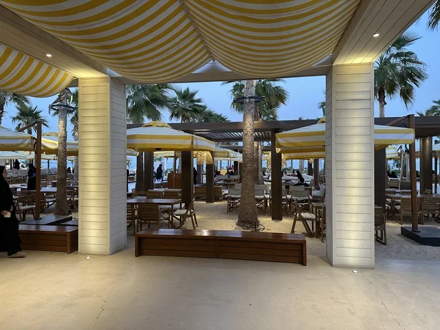 تصميم خارجي بأسقف قماشية مخططة باللونين الأصفر والأبيض، سولت البحر وجهة ترفيهية وشاطئية عائلية تقع على طريق الكورنيش في الخبر في المملكة العربية السعودية، تجديدات إضافية وتطويرات في 3  يناير 2026، شاطئ رملي مع وجود مظلات وأشجار نخيل