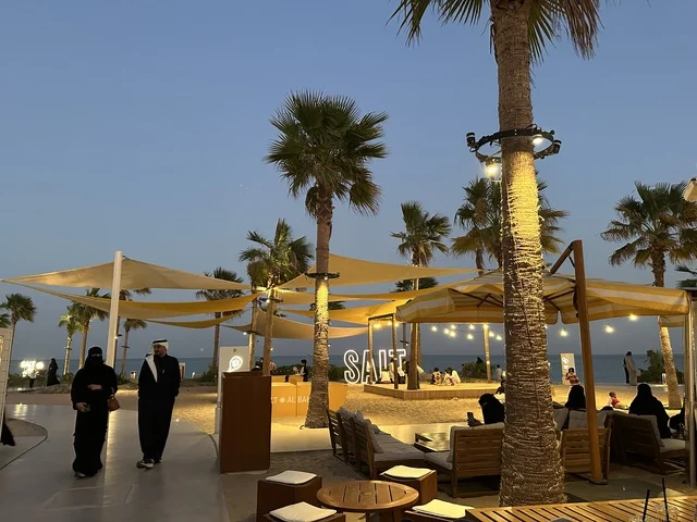 منطقة جلوس خارجية ساحلية في المساء ، سولت البحر وجهة ترفيهية وشاطئية عائلية تقع على طريق الكورنيش في الخبر في المملكة العربية السعودية، تجديدات إضافية وتطويرات في 3  يناير 2026، شاطئ رملي مع وجود مظلات وأشجار نخيل