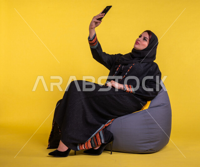 بورتريه لسيدة عربية خليجية سعودية مبتسمة تستخدم الهاتف المحمول لاتقاط صورة سيلفي ، صورة تذكارية ، اجراء مكالمات الفيديو، اجتماع عمل عن بعد ، التواصل مع الاصدقاء ،  التسلية والترفيه ،  خلفية صفراء