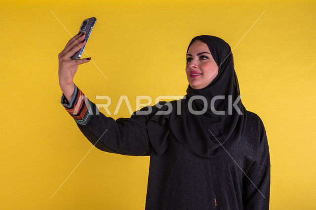 بورتريه لسيدة عربية خليجية سعودية مبتسمة تستخدم الهاتف المحمول لاتقاط صورة سيلفي ، صورة تذكارية ، اجراء مكالمات الفيديو، اجتماع عمل عن بعد ، التواصل مع الاصدقاء ،  التسلية والترفيه ،  خلفية صفراء