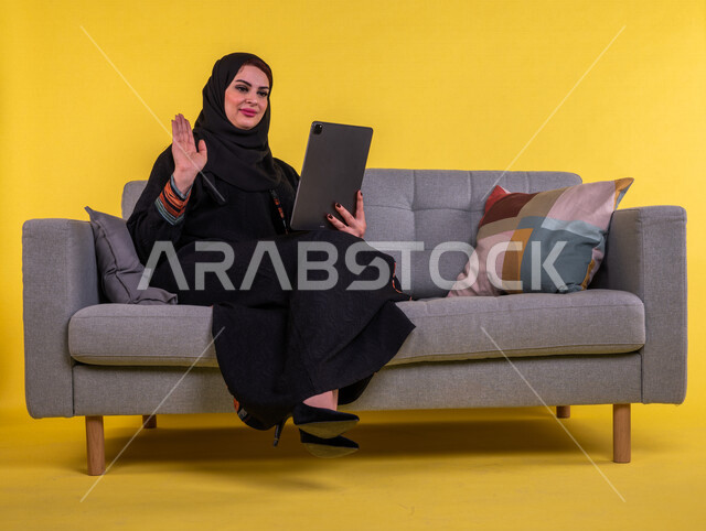 بورتريه لسيدة عربية خليجية سعودية مبتسمة تستخدم جهاز التابلت لاجراء مكالمة فيديو ، ترفع يدها لالقاء التحية والسلام ،   اجتماع عمل عن بعد ، التواصل مع الاصدقاء ،  التسلية والترفيه ،  خلفية صفراء