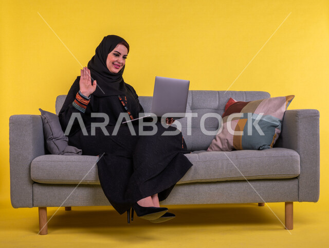 بورتريه لسيدة عربية خليجية سعودية مبتسمة تستخدم جهاز الحاسوب المحمول لاجراء مكالمة فيديو ، ترفع يدها لالقاء التحية والسلام ،   اجتماع عمل عن بعد ، التواصل مع الاصدقاء ،  التسلية والترفيه ،  خلفية صفراء