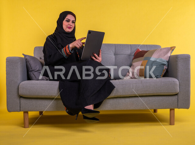 سيدة عربية خليجية سعودية مبتسمة تستخدم جهاز التابلت لإجراء مكالمة فيديو ، تصفح الإنترنت , اجتماع عمل عن بعد ، التواصل مع الاصدقاء ، التسلية والترفيه ، خلفية صفراء .