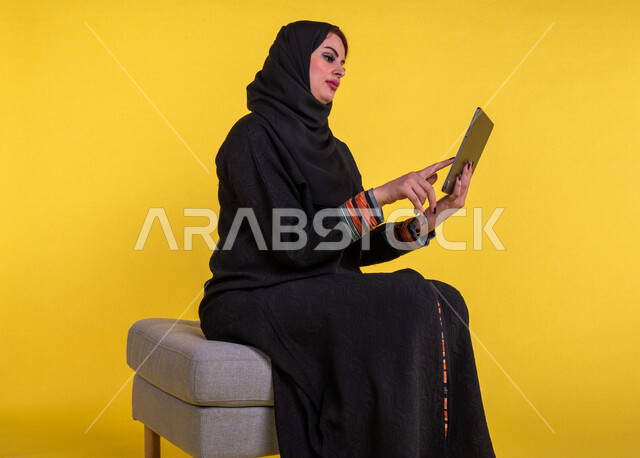 سيدة عربية خليجية سعودية مبتسمة تستخدم جهاز التابلت لإجراء مكالمة فيديو ، تصفح الإنترنت , اجتماع عمل عن بعد ، التواصل مع الاصدقاء ، التسلية والترفيه ، خلفية صفراء .