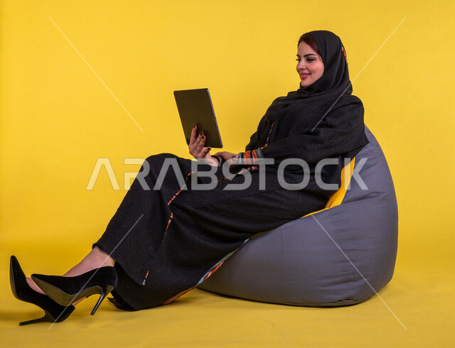 سيدة عربية خليجية سعودية مبتسمة تستخدم جهاز التابلت لإجراء مكالمة فيديو ، تصفح الإنترنت , اجتماع عمل عن بعد ، التواصل مع الاصدقاء ، التسلية والترفيه ، خلفية صفراء .