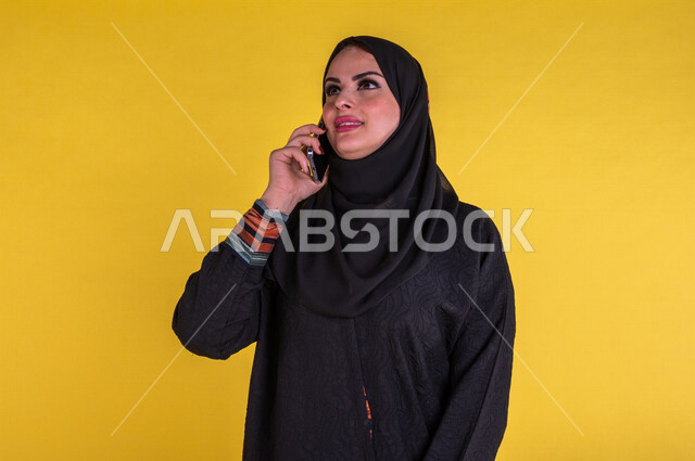 سيدة عربية خليجية سعودية مبتسمة تستخدم الهاتف المحمول للتواصل ، اجراء مكالمات عبر الهاتف النقال ، التواصل مع الاصدقاء ، التسلية والترفيه ، خلفية صفراء .