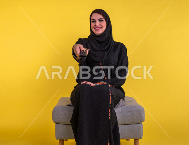 بورتريه امرأة عربية سعودية خليجية تمسك جهاز التحكم عن بعد بالتلفاز , مشاهدة الأفلام والمسلسلات , البرامج الثقافية والأخبار , قضاء وقت الفراغ , التسلية والترفيه , خلفية صفراء .