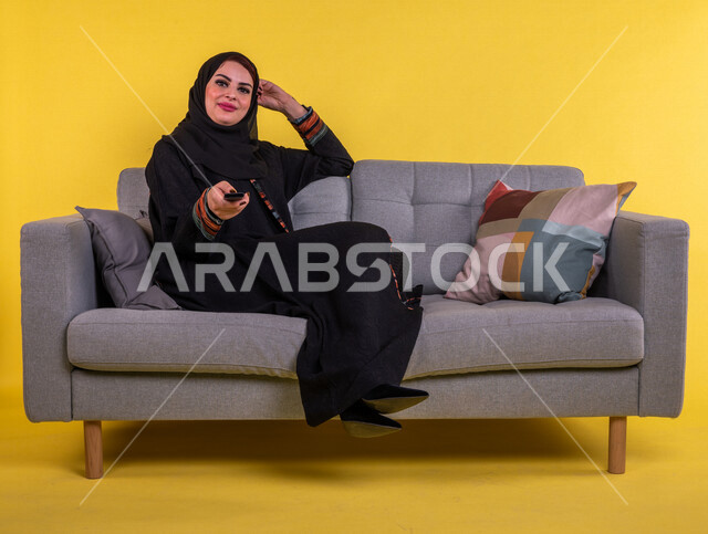 بورتريه امرأة عربية سعودية خليجية تمسك جهاز التحكم عن بعد بالتلفاز , مشاهدة الأفلام والمسلسلات , البرامج الثقافية والأخبار , قضاء وقت الفراغ , التسلية والترفيه , خلفية صفراء .