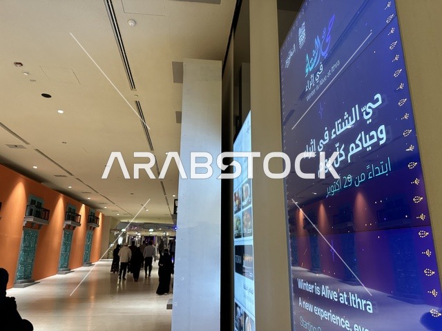 An internal corridor in a cultural center, a blue digital display screen, visitors wandering inside the center, internal digital directional signs, a modern internal cultural environment, an interior image of the Ithra Center in the Dhahran area of Saudi Arabia, the King Abdulaziz Center for World Culture.