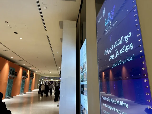 An internal corridor in a cultural center, a blue digital display screen, visitors wandering inside the center, internal digital directional signs, a modern internal cultural environment, an interior image of the Ithra Center in the Dhahran area of Saudi Arabia, the King Abdulaziz Center for World Culture.