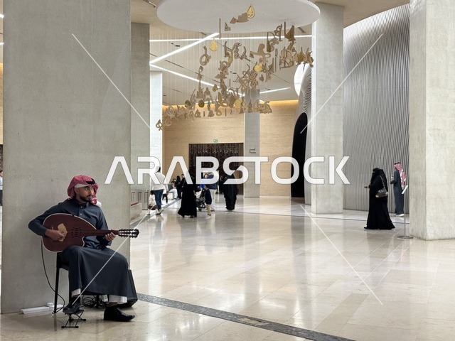 رجل عربي خليجي سعودي يرتدي الثوب التقليدي والشماغ يعزف على آلة العود داخل مركز إثراء الثقافي بالرياض، أنشطة فنية داخل مبنى ثقافي حديث، تجول السياح و الزوار فى الممر الداخلي