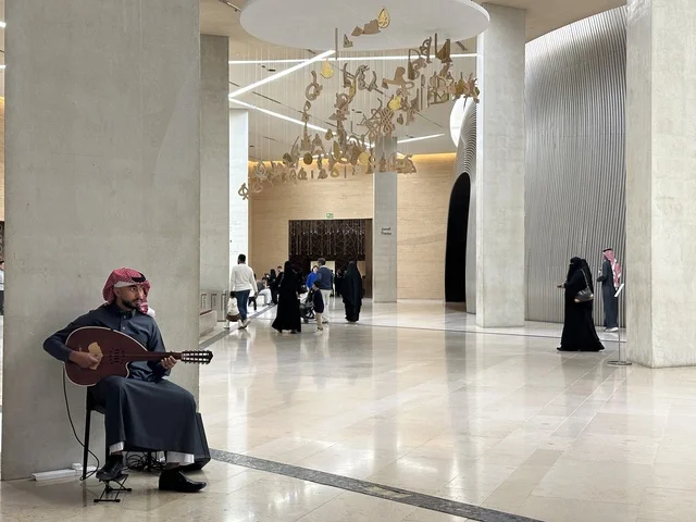 رجل عربي خليجي سعودي يرتدي الثوب التقليدي والشماغ يعزف على آلة العود داخل مركز إثراء الثقافي بالرياض، أنشطة فنية داخل مبنى ثقافي حديث، تجول السياح و الزوار فى الممر الداخلي